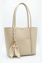 A light beige leather tote bag with dual top handles, a front slip pocket, and a decorative braided handle accent tied with a matching beige and pink striped scarf.